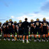 The varsity football team awaits on the sidelines watching their team members play.